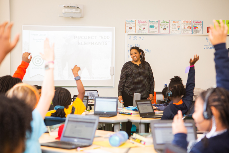 Teacher stands in classroom with students raising their hands