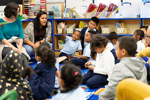 Two teachers work with a group of young students, sitting on the floor in a classroom