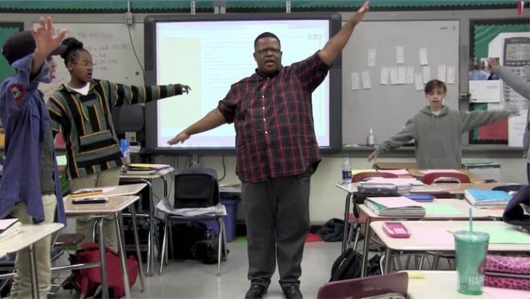 man stands in front of classroom with his arms up