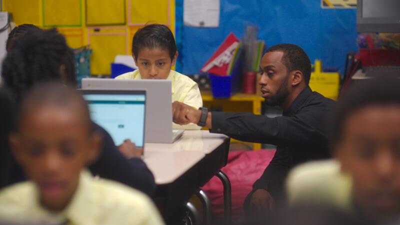 teacher talks with student working on laptop in classroom