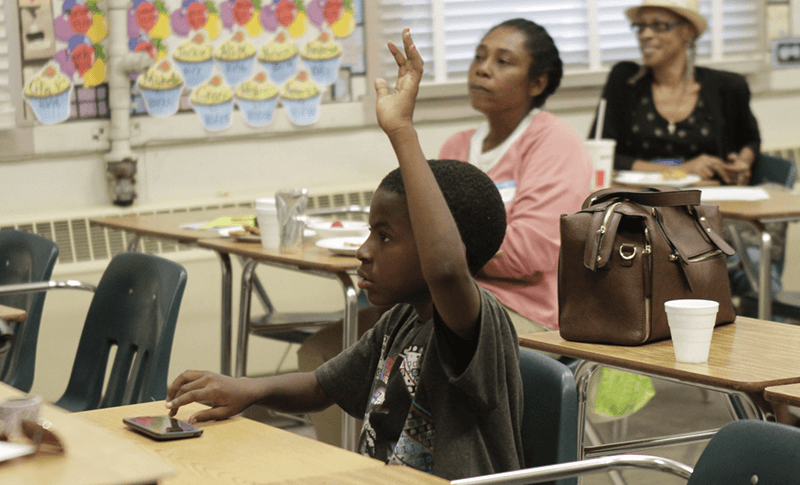 student raises his hand while sitting at a classroom desk
