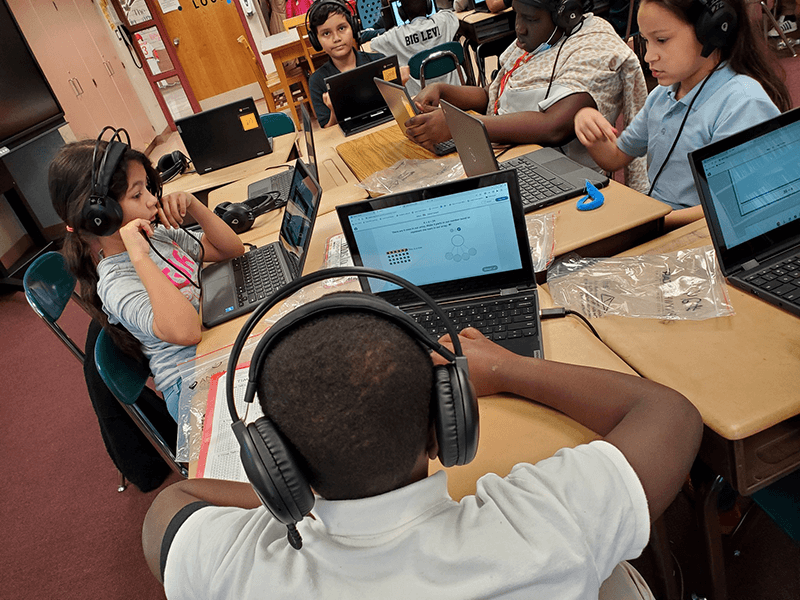 Students sit around classroom desks working on laptops wit headphones on
