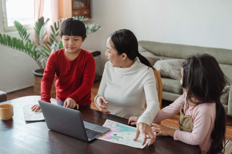 parent sits with two children at a table with laptop
